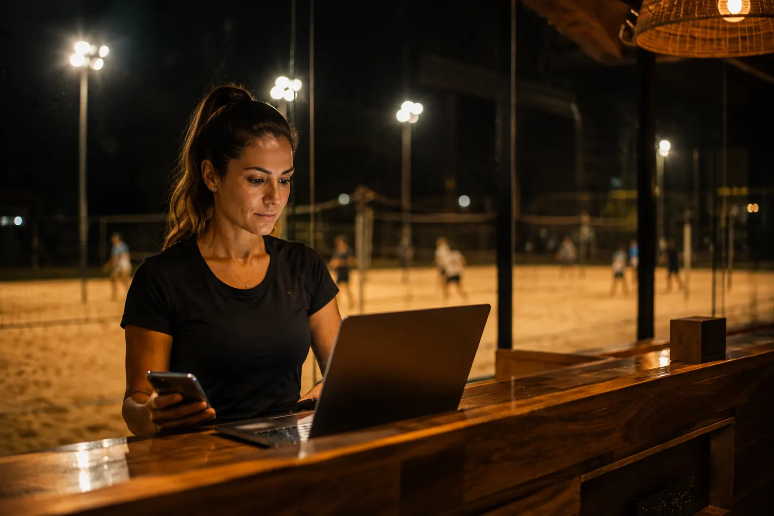 Gestora na recepção de uma arena consultando o notebook e o celular, com quadras de areia iluminadas ao fundo